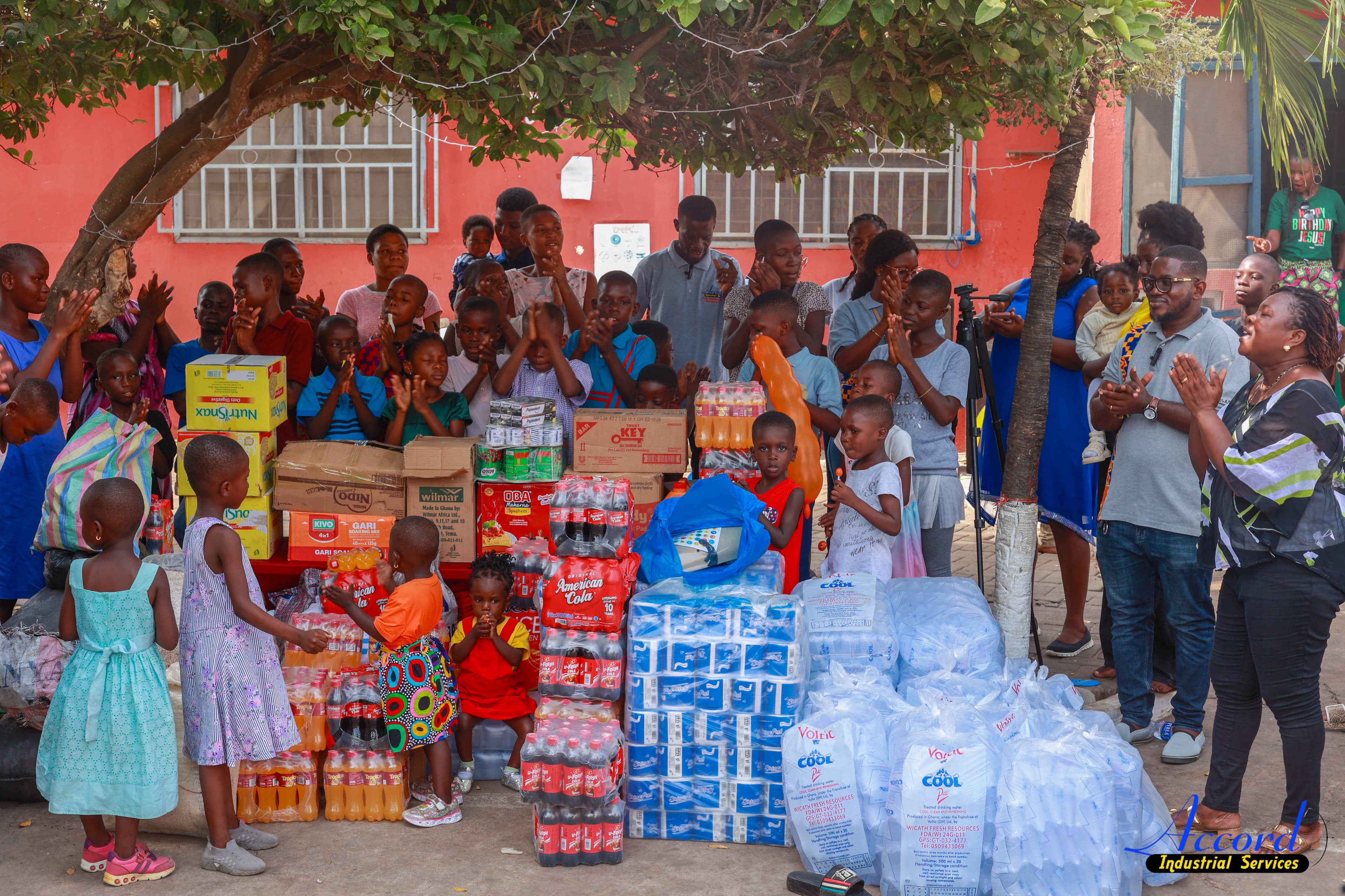 Donated items including water, food, and supplies stacked with children gathered around